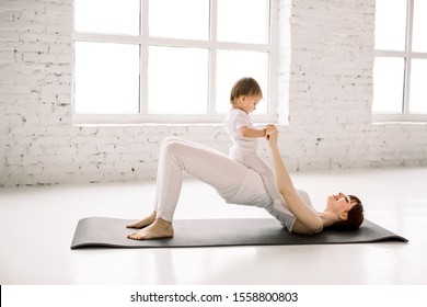 Young happy mother working out, doing butt bridge exercise, wearing white sportswear, little baby girl on her tummy, fitness, postnatal yoga. Healthy lifestyle concept
