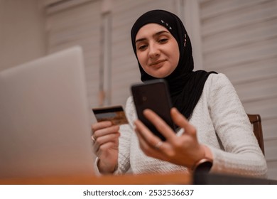 A young happy Muslim woman using mobile phone and holding credit card checking balance account or making money transfer. Online Payment Internet Banking Concept. - Powered by Shutterstock