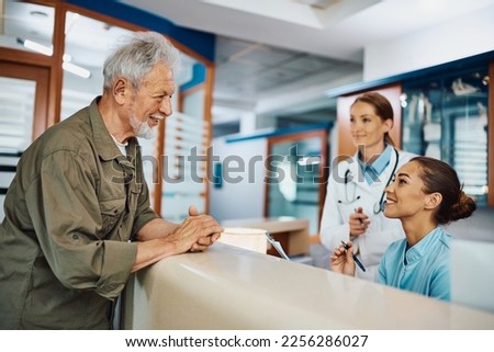 Young happy nurse taking notes while communicating with mature patient at reception desk at medical clinic.
