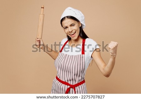 Young happy overjoyed housewife housekeeper chef baker latin woman wear striped apron toque hat hold rolling pink do winer gesture isolated on plain pastel light beige background. Cook food concept