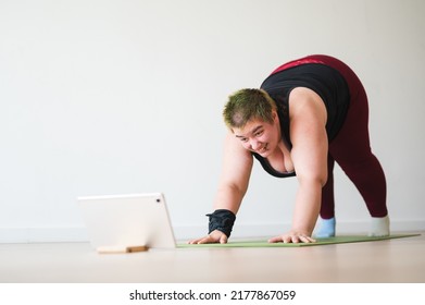 Young Happy Overweight Woman In Sportswear Stretching On Yoga Mat And Follow Exercise Instruction On Mobile Device Tablet. Home Workout For Obese Person Concept. Yoga Asana Down Dog Plus Size Female. 