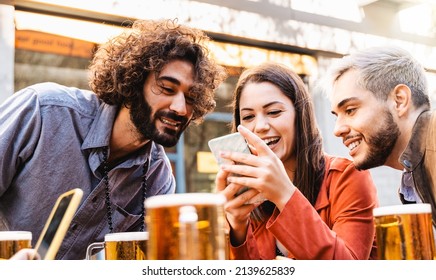 Young happy people having fun using mobile phone while drinking beer at brewery bar outdoor - Focus on center girl face