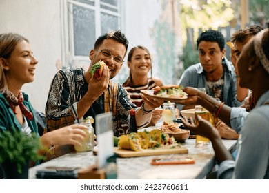 Young happy people having lunch together. Focus is on man passing tacos to his female friend. 