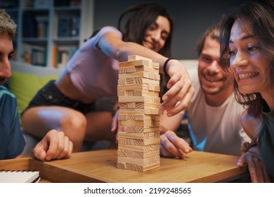 Young happy people playing Jenga at home, a girl is removing a wooden block from the tower and the others are watching - Powered by Shutterstock