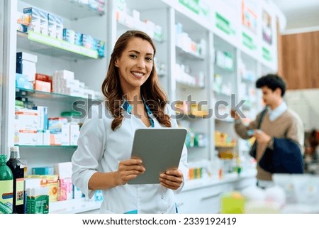 Young happy pharmacist working on digital tablet in drugstore and looking at camera. Her customer is in the background.