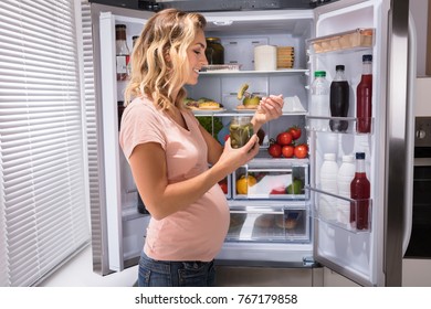 Young Happy Pregnant Woman Eating Pickle From Jar Near Refrigerator