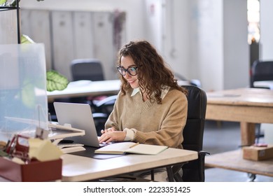 Young happy professional business woman employee sitting at desk working on laptop in modern corporate office interior. Smiling female worker using computer technology typing browsing web.