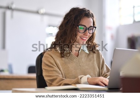 Young happy professional business woman worker employee sitting at desk working on laptop in corporate office. Smiling female student using computer technology learning online, doing web research.