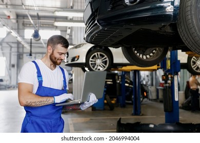 Young happy professional technician mechanic man in denim blue overalls t-shirt use hold laptop pc computer stand near car lift check technical condition work in vehicle repair shop workshop indoors