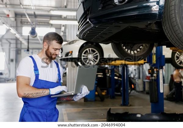 Young happy professional technician mechanic man\
in denim blue overalls t-shirt use hold laptop pc computer stand\
near car lift check technical condition work in vehicle repair shop\
workshop indoors