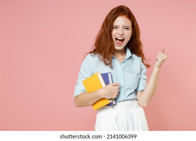 Young happy redhead student woman in blue shirt hold book do winner gesture clench fist celebrate isolated on pastel pink background studio portrait. Education high school university college concept.