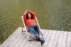 Young Happy Relaxed African American Woman With Curly Hair In Orange Warm Sweater Relaxing Resting On Lounge Chair Near Pond Or River On Autumn Weekend Day. People And Recreation In Nature