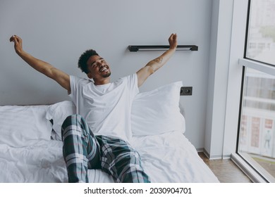 Young happy rested african american man in nightwerar do morning hand stretching lying in bed rest relax spend time in bedroom lounge home in own room house wake up dream be lost in reverie good day.