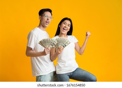 Young Happy Rich Asian Couple Holding Bunch Of Money Over Yellow Studio Background