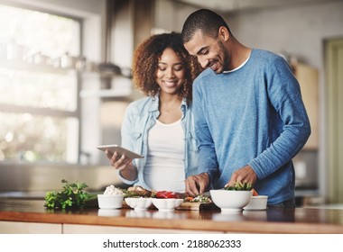 Young, Happy And Romantic Couple Cooking Healthy Food Together Following Recipes Online On A Tablet. Smiling Husband And Wife Making Dinner Or Lunch In The Kitchen At Home