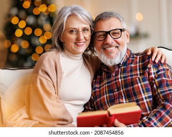 Young happy senior family couple in love enjoying winter holidays together at home, smiling elderly man and woman read book and embrace while relaxing on cozy sofa, spending leisure time on Christmas - Powered by Shutterstock