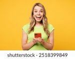 Young happy shocked surprised blonde woman she wears green t-shirt casual clothes hold in hand use mobile cell phone chat online isolated on plain yellow orange background studio. Lifestyle concept