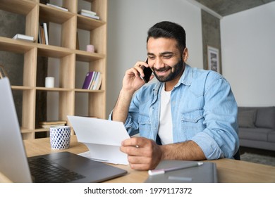 Young Happy Smiling Adult Indian Hispanic Businessman Talking On Mobile Cellphone Arranging Meeting With Employee Reading Paper CV Sitting At Desk At Home Office. Remote Recruitment Work Concept.