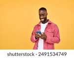 Young happy smiling African American Black hipster man user customer with cellphone in hands standing isolated on yellow background holding smartphone using mobile cell phone scrolling social media.