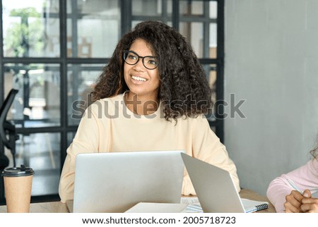 Young happy smiling African American girl student in eyeglasses at desk working using pc laptop computer at modern office university college campus listening lesson class. Education concept.