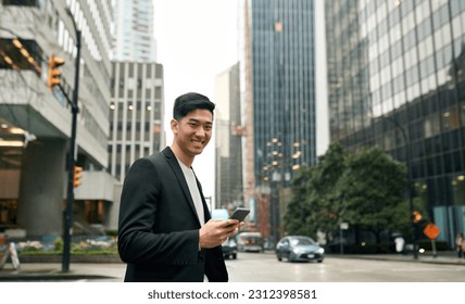 Young happy smiling Asian business man professional manager with cellphone in hands standing on big city street holding mobile cell phone, using smartphone apps, looking at camera, portrait. - Shutterstock ID 2312398581