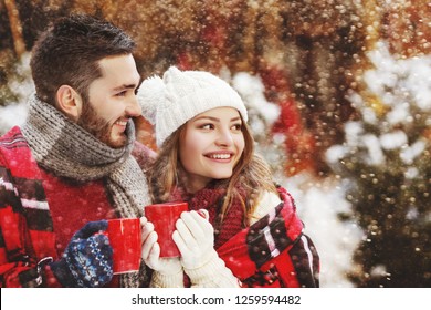 Young happy smiling couple bundle up plaid, models holding red mugs with hot drink, looking aside, posing in street. Snowfall. Christmas celebration, winter holidays concept. Copy, empty space 