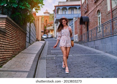 Young happy smiling cute beautiful brunette traveler girl with long legs wearing hat, round straw bag and short jumpsuit walking alone around the city at sunny summer day