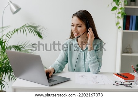 Young happy smiling employee operator business woman in set microphone headset blue shirt for helpline assistance sit work at call center workplace desk with laptop pc computer typing office indoors
