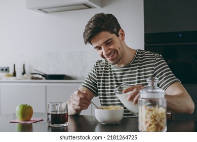 Young Happy Smiling Man 20s Wear Striped T-shirt Eat Breakfast Muesli Cereals With Fruit In Bowl Pouring Milk Sit By Table Cooking Food In Light Kitchen At Home Alone Healthy Diet Lifestyle Concept.