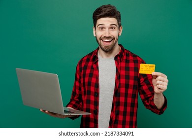 Young Happy Smiling Man He 20s In Red Shirt Grey T-shirt Using Laptop Pc Computer Hold Credit Bank Card Doing Online Shopping Order Delivery Booking Tour Isolated On Plain Dark Green Background Studio