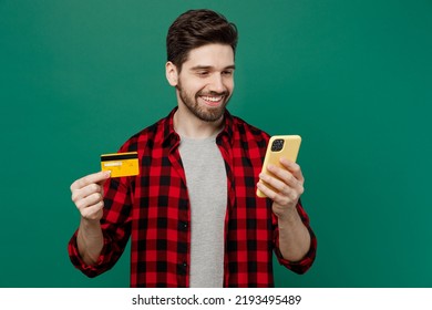 Young Happy Smiling Man He 20s Wearing Red Shirt Grey T-shirt Using Mobile Cell Phone Hold Credit Bank Card Doing Online Shopping Order Delivery Booking Tour Isolated On Plain Dark Green Background