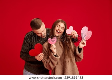 Young happy smiling man and woman, couple, boyfriend and girlfriend posing with paper hearts against red studio background. Concept of love, relationship, Valentine's Day, emotions, lifestyle