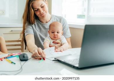 A young happy smiling mother with a baby in her arms sits at the table and works on a laptop and with documents.