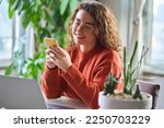 Young happy smiling pretty woman sitting at table holding smartphone using cellphone modern technology, looking at mobile phone while remote working or learning, texting messages at home.