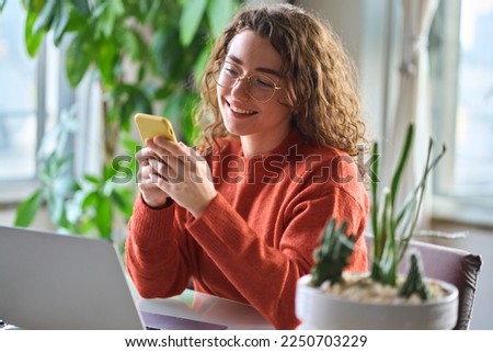 Young happy smiling pretty woman sitting at table holding smartphone using cellphone modern technology, looking at mobile phone while remote working or learning, texting messages at home.