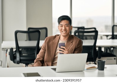 Young happy smiling professional Asian Japanese business man manager executive holding smartphone using mobile cell phone looking at camera working in office with digital cellular technology. - Powered by Shutterstock