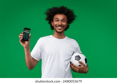 Young happy smiling soccer supporter man showing smartphone with bookmaker's mobile application interface on its screen to the camera. His bet played. Isolated over green background.