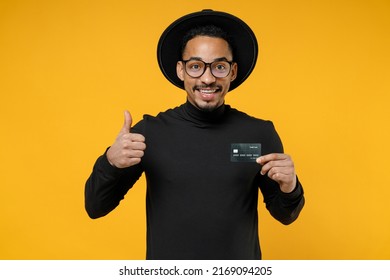 Young Happy Smiling Successful American African Man 20s In Stylish Black Shirt Hat Eyeglasses Holding Credit Bank Card Show Thumb Up Like Gesture Isolated On Yellow Orange Background Studio Portrait.