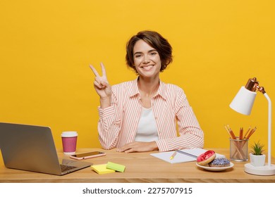 Young happy smiling successful employee business woman wear casual shirt sit work at office desk with pc laptop show v-sign gesture isolated on plain yellow color background Achievement career concept