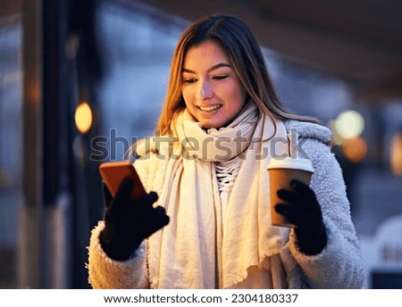 Young happy smiling teenager girl walking on a city street in jacket on a cold winter evening with takeaway coffee in her hand while browsing on the internet on her mobile phone