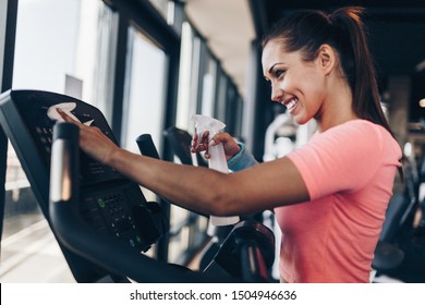 Young Happy And Smiling Woman Cleaning And Weeping Expensive Fitness Gym Equipment With Sprayer And Cloth.