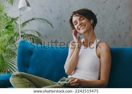 Young happy smiling woman wear white tank shirt earphones listen music hold use mobile cell phone sit on blue sofa stay at home flat rest relax spend free spare time in living room indoors grey wall