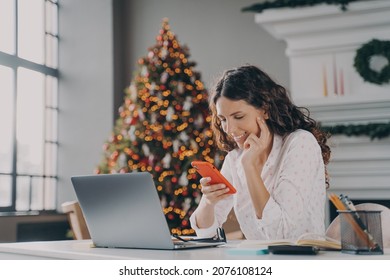 Young happy spanish businesswoman using smartphone while working laptop at home on Christmas, sitting at desk against fir xmas tree with lights, reading sms message on mobile during winter holidays - Powered by Shutterstock