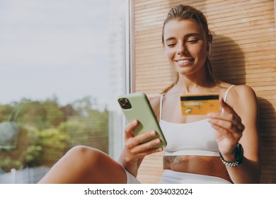 Young happy sportswoman woman in white sportswear earphones listen music use mobile cell phone credit bank card shopping online order delivery book tour pay gym membership sit on windowsill indoor.