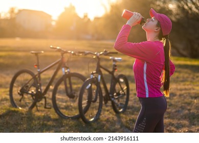 Young happy sporty female wearing rose clothes drinks from water bottle with two bicycles in the background at sunset - Shutterstock ID 2143916111