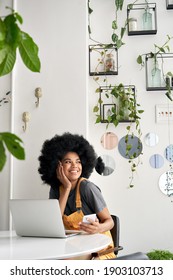 Young happy stylish African American gen z hipster lady student with Afro hair laughing, looking through window, sitting at table in modern cozy cafe interior, holding phone, using laptop.