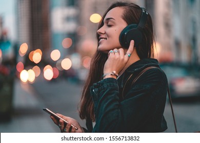 Young happy stylish trendy casual hipster woman teenager listening to music on a black wireless headphone while walking around the city. Music lover enjoying music