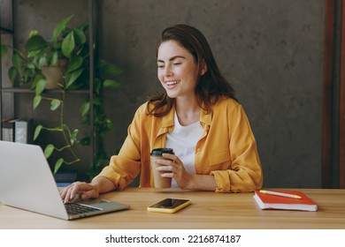 Young happy successful employee business woman wear casual yellow shirt hold brown cup coffee hold use on laptop pc computer sit work at wooden office desk with pc laptop. Achievement career concept