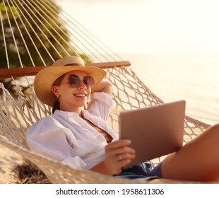 Young happy successful woman, smiling female freelancer in straw hat and sunglasses using digital tablet while lying and relaxing in the hammock on tropical sandy beach on hot sunny day. Distance work - Powered by Shutterstock