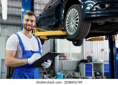 Young Happy Technician Mechanic Man Wears Denim Overalls Use Hold Clipboard Papers Document Writing Estimate Stand Near Car Lift Check Technical Condition Work In Vehicle Repair Shop Workshop Indoors